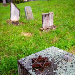 Rusty Star of David on a symbolic memorial at an old Jewish cemetery with weathered tombstones. Historic site surrounded by green grass and moss.