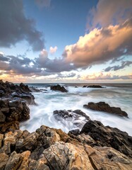 Obraz premium Dramatic coastal scene at sunset. Ocean waves crash against dark and light rocks. Pastel clouds fill the sky