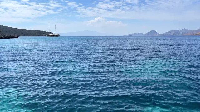 A stunning view of deep blue sea with gentle waves, a traditional gulet near a rocky coastline, and hazy mountains in the distance under a partly cloudy sky.