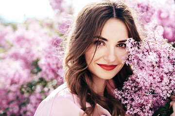 Fototapeta premium Young beautiful smiling woman. Sexy carefree model posing in the street. Positive model near blooming flower lilac bush beaming. Summertime, pink colours. Closeup beauty portrait, red lips