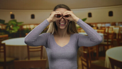 Woman smiling playfully while mimicking binoculars with hands in a cozy restaurant setting,...