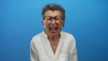 Woman expressing emotions with closed eyes and open mouth against solid blue background wearing white blouse and glasses.