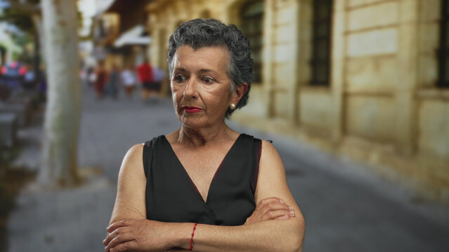Senior woman with grey hair crosses arms and looks serious on an old town street, highlighting urban life and architectural charm.