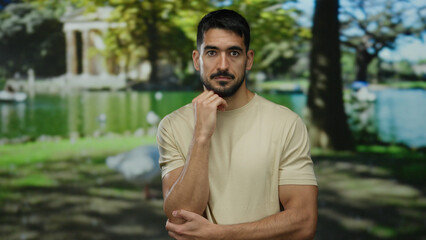 Pensive man outdoors in park setting with thoughtful expression wearing casual beige shirt surrounded by lush trees and peaceful lake under vibrant sunlight