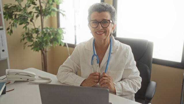 Senior woman doctor with grey hair in a clinic office holding a stethoscope smiling at her desk surrounded by office equipment and a plant.