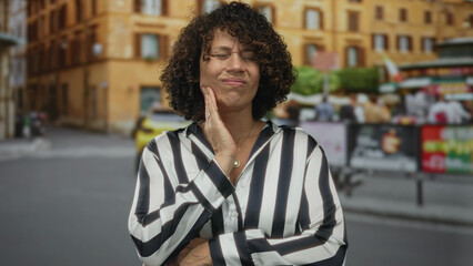 Woman pressing cheek with hand while wincing on a sunlit urban street with blurred building behind;...