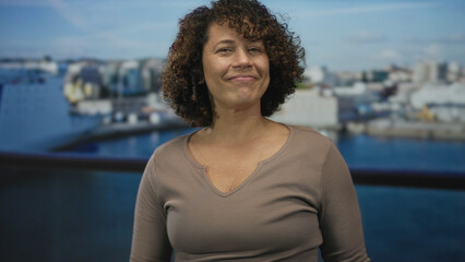 Hispanic woman flexes bare bicep aboard a boat deck at port in daylight, smiling with closed eyes;...