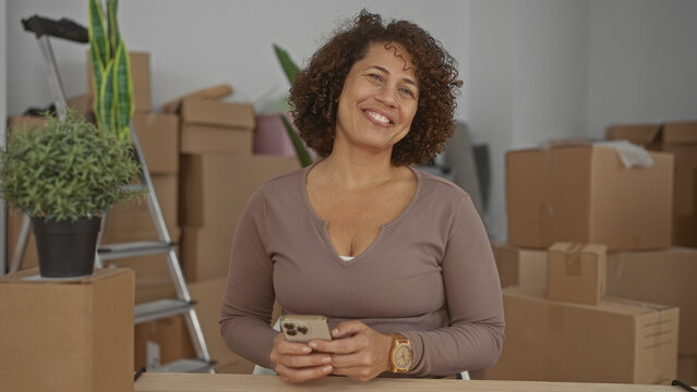Woman holds smartphone texting inside building surrounded by moving boxes and a ladder; anticipation and hope.