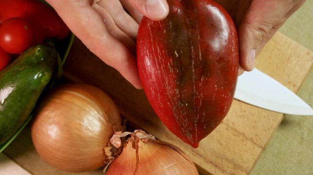 Male hands hold fresh bell pepper on wooden board with onion, tomatoes and knife. Bell pepper shows vivid color and texture, pepper near vegetables highlights beauty. Concept of healthy homemade food.
