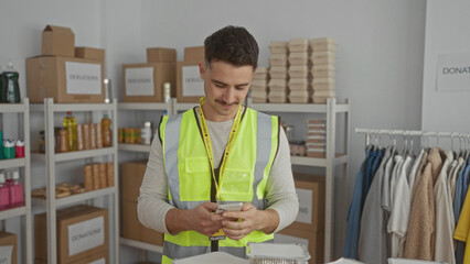 Young hispanic man in reflective vest takes a selfie at an indoor charity center filled with donations, surrounded by organized boxes and clothing racks.