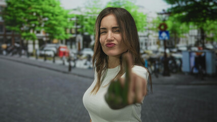 Woman flips middle finger on city street with blurred buildings under bright daylight; defiance.