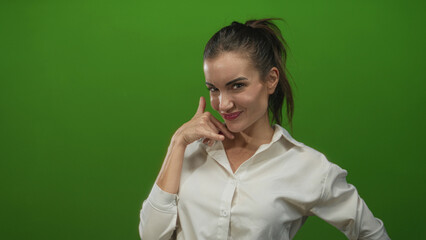 Young hispanic woman points finger toward camera and makes phone gesture against vivid green studio set; playful charm.