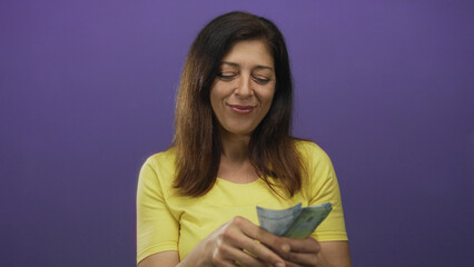 Middle age hispanic woman confidently counts chilean peso banknotes in studio against purple wall; contentment.