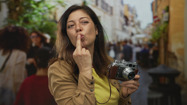 Hispanic middle age woman holding vintage camera shows middle finger with a grin on busy street; defiance.