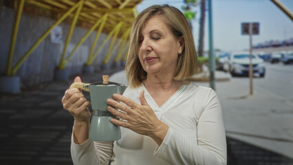 Caucasian middle aged woman holds green moka pot while covering mouth in yawn on urban street under...