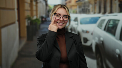 Fototapeta premium Woman pointing at eye with smile on city street wearing glasses and blazer, with cars in background, capturing casual urban setting and friendly gesture.