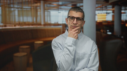 Man thinking deeply in a modern coffee shop indoors, wearing glasses and a striped shirt, with a thoughtful expression in a relaxed restaurant setting.