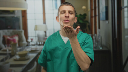 Caucasian male doctor in green uniform stands indoors blowing a kiss in a warmly lit room,...