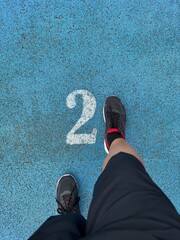 Man standing on blue running track with number two marking, top view of legs and shoes, sports and fitness concept