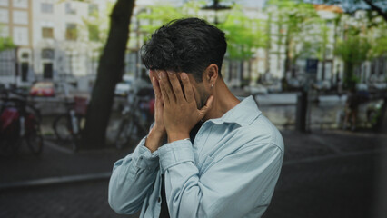 Man covers face with both hands in denim jacket on crowded urban street beside canal at dusk; anxiety.