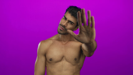 Young hispanic man with firm expression pushing palm forward across bare chest in purple studio;...