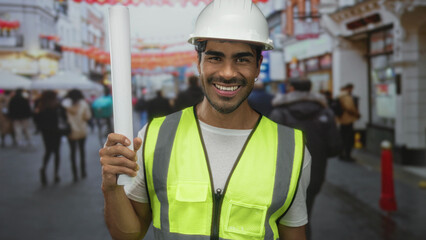 Man wearing a hardhat examines rolled document on a crowded city street during daytime; determination.