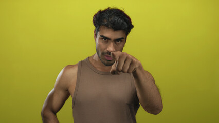 Fototapeta premium Young hispanic man in a brown tank top with a bare muscular arm points finger directly at camera in studio; confidence.