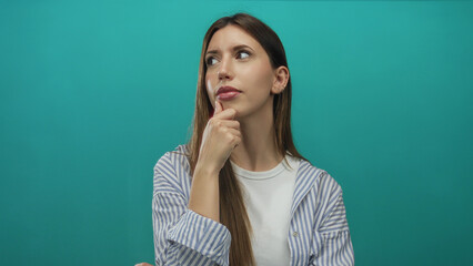 Young hispanic woman wearing striped shirt with hand on chin in teal studio scene; contemplation...