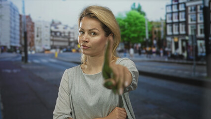 Blonde woman points her finger forward at busy city street intersection wearing gray blouse; bold confidence.
