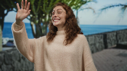 Woman smiling and waving on a sunny seaside promenade wearing a cozy sweater with a vibrant...