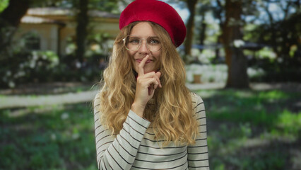 Woman places finger against lips in a shushing pose inside a quiet park beside a blonde young model...