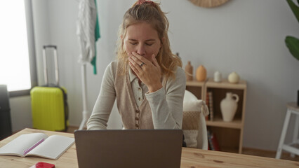 Woman covers mouth while yawning at a laptop, her blonde young silhouette brightening a home office with a desk in an indoor workspace.