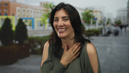 Middle age hispanic woman laughing with hand on cheek on sunlit bustling street; warmth joy positivity.