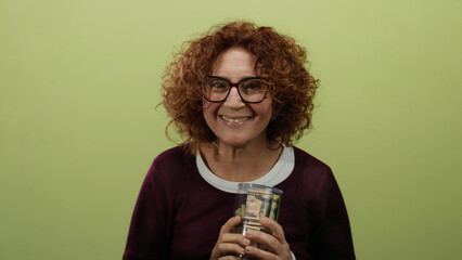 Woman holding a clear bottle with us dollar bills against a green background, smiling with curly...