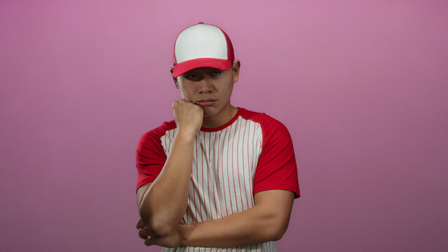 Young man in a red baseball uniform appears deep in thought against an isolated pink wall background.