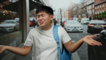 Young man with blue backpack shrugs on a bustling city street, conveying uncertainty or hesitation...