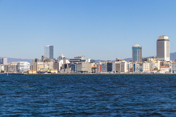 Obraz premium Modern city skyline along the coastline with sea and mountains in the background on a clear sunny day.
