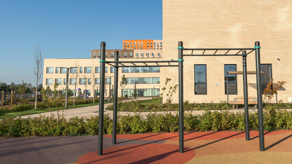 Modern school building exterior with outdoor sports area featuring metal pull-up bars and colorful rubber flooring under clear blue sky, representing education, healthy lifestyle, urban development