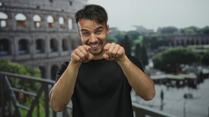 Young hispanic man gesturing at roman colosseum, smiling in black shirt with cityscape of iconic...