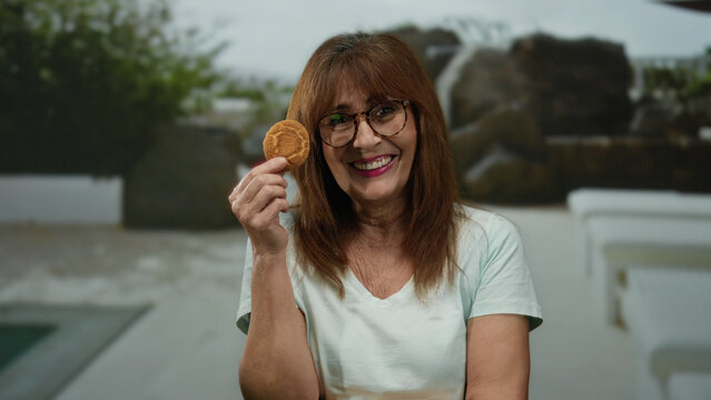 Hispanic senior woman smiling with cookie at resort poolside capturing a joyful outdoor vacation moment. - Powered by Adobe