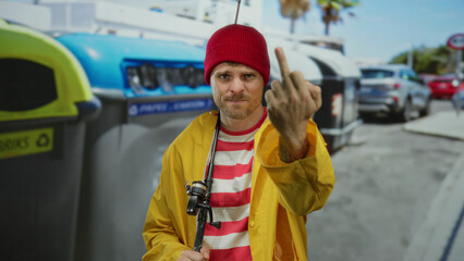 Young man in yellow jacket and red beanie with fishing rod makes rude gesture on city street with...
