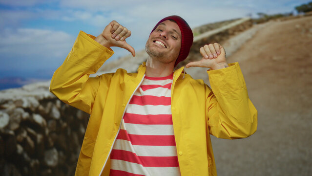 Young man in yellow raincoat and red striped shirt proudly points at himself on a sunny urban street with a mountain landscape background.