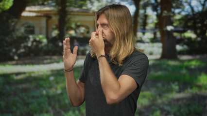 Man with long blond hair pinches nose and raises open palm to sniff something in green park under...
