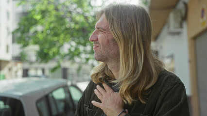 Man with long blond hair holds cheeks with both hands in green jacket on sunlit urban street beside...