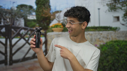 Man wearing round glasses points black electric shaver at building gate outdoors; technology...