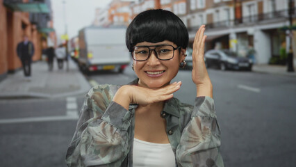 Fototapeta premium Chinese woman smiling on urban street poses with glasses and hands framing face, capturing outdoor city vibe.