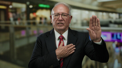Senior man in a suit standing in a mall with one hand on his chest and the other raised, conveying...