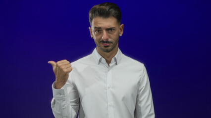 Man pointing bare thumb to left in blue studio setting under bright even lighting; self doubt hesitation uncertainty.