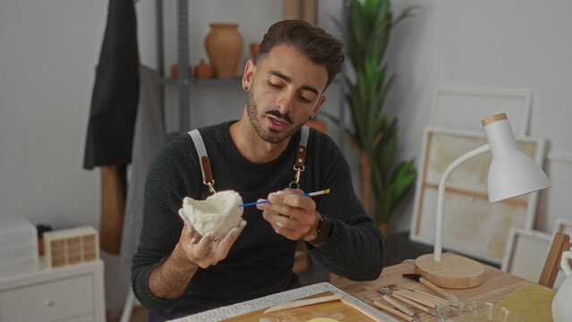 Man holding clay bowl with hand while painting it with brush over wooden table in studio; creative focus.