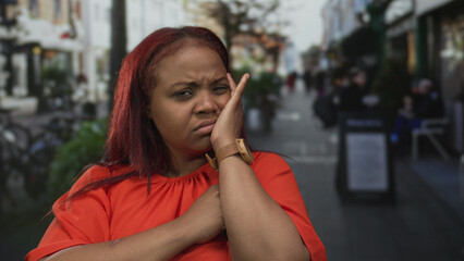 Woman holds hand to cheek on busy street in urban scene with blurred storefronts and passersby; indignation.